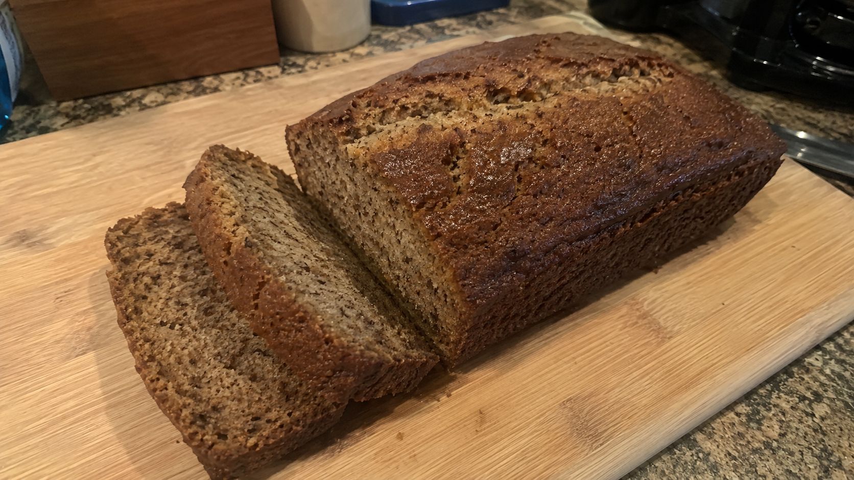 A golden-brown loaf of homemade banana bread sliced on a wooden cutting board, showing a moist tender crumb, with ripe bananas in the background