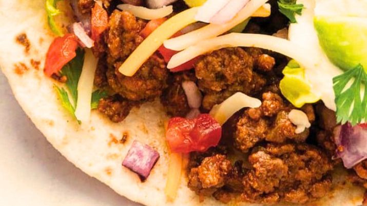 Seasoned ground beef tacos in warm corn tortillas topped with shredded cheese, diced tomato, fresh cilantro, and a squeeze of lime on a wooden board