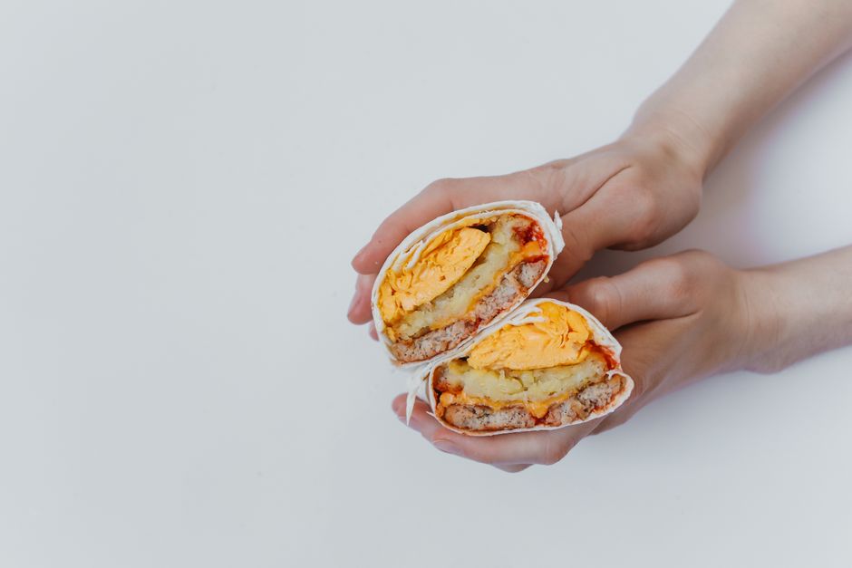 Three golden toasted breakfast burritos on a wooden cutting board, sliced in half to reveal layers of scrambled eggs, crispy potatoes, sausage, and melted cheddar cheese, with salsa and hot sauce on the side