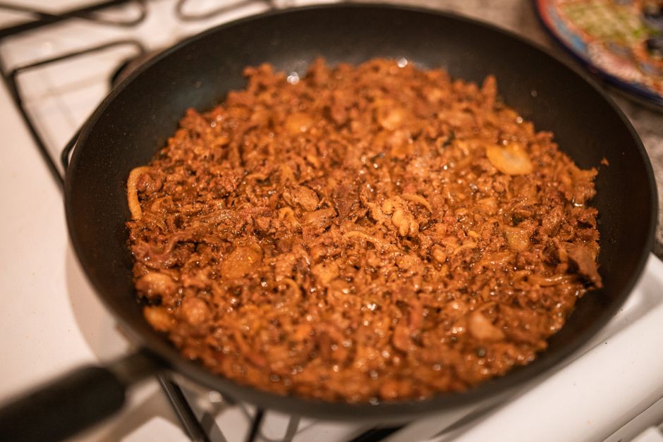 Thinly sliced carne asada on a wooden cutting board with charred edges, surrounded by warm corn tortillas, diced white onion, fresh cilantro, lime wedges, and red salsa