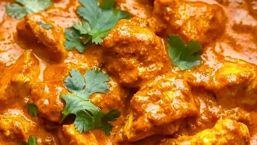 A bowl of chicken tikka masala with tender charred chicken pieces in a deep orange-red creamy sauce, garnished with fresh cilantro, served alongside basmati rice and warm naan bread