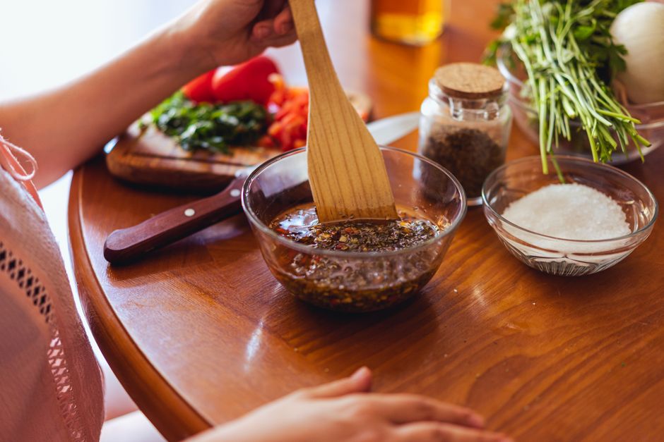 A small white bowl filled with vibrant green chimichurri sauce, chunky with fresh parsley and flecked with red pepper, resting on a dark wooden board beside a sliced skirt steak