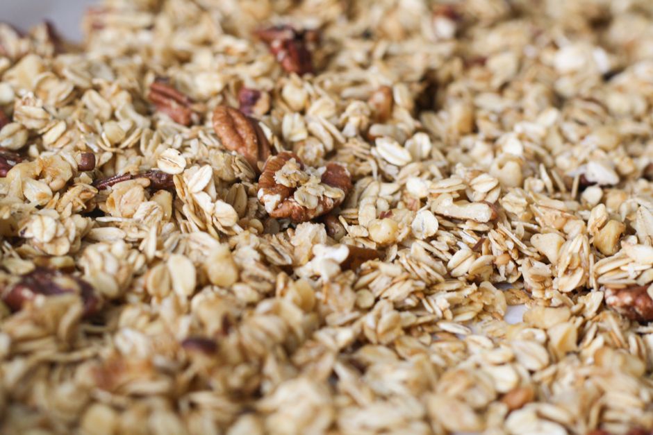 A wide-mouth mason jar filled with golden homemade granola clusters, surrounded by scattered oats, dried cranberries, and pecans on a white linen surface