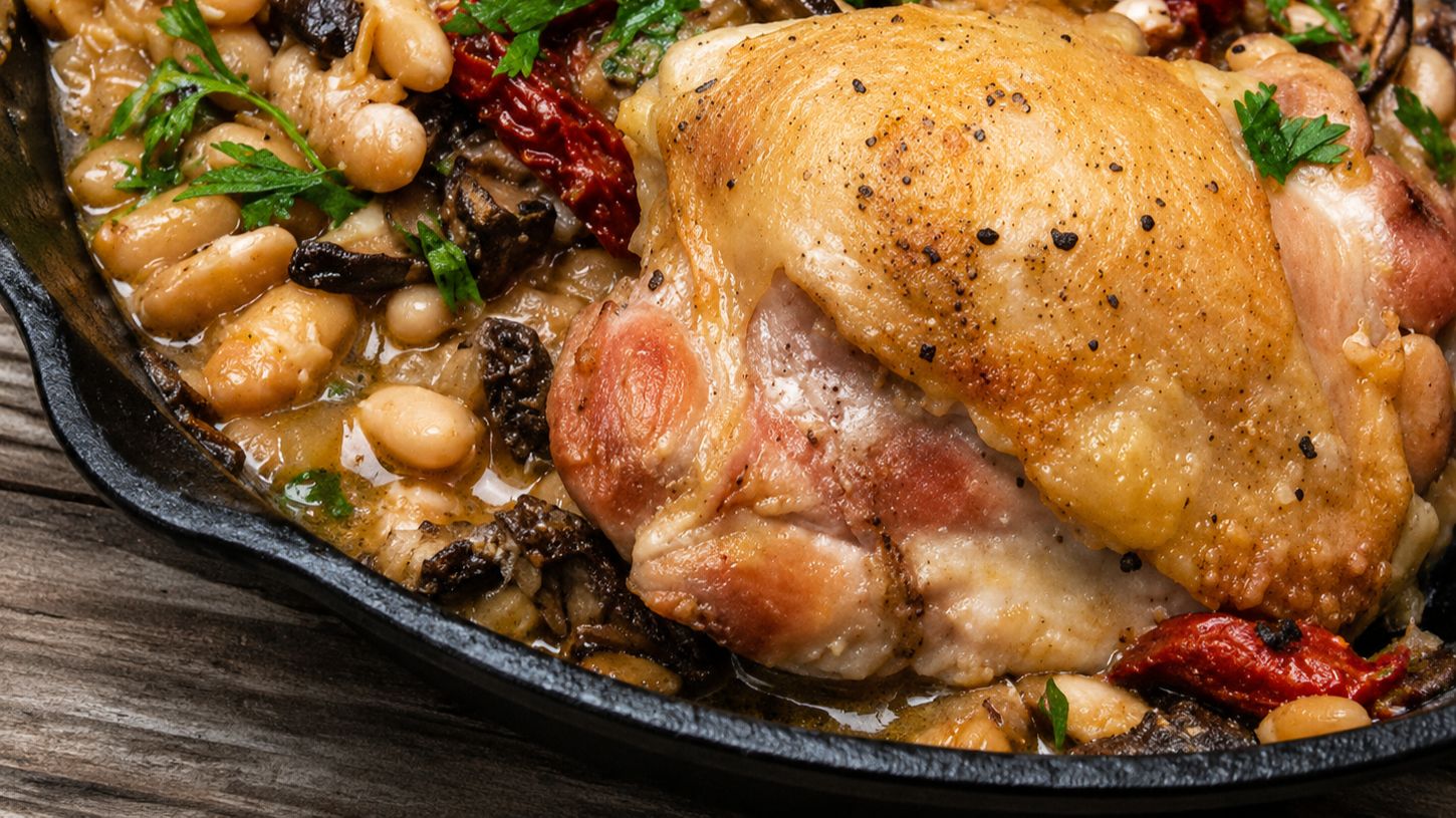 Skillet chicken and white beans with sun-dried tomatoes, wilted spinach, and Parmesan cheese in a cast iron pan, garnished with fresh parsley