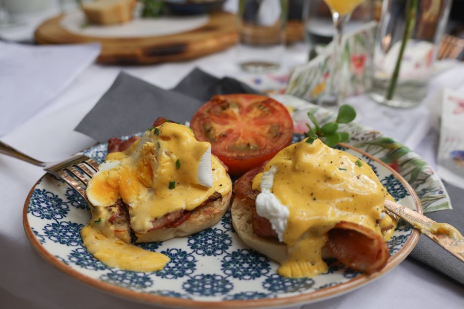 A small white pitcher pouring glossy golden hollandaise sauce over poached eggs on an English muffin, with steam rising and a sprig of fresh chervil on the plate