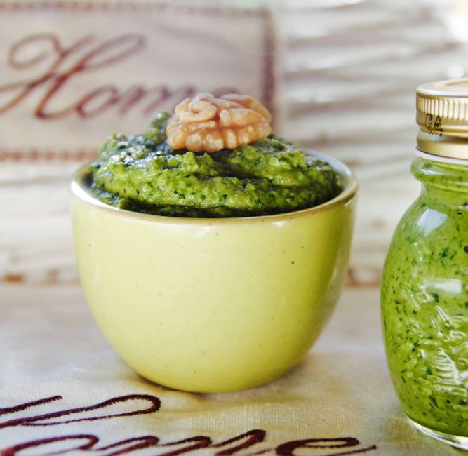 A small glass jar of vibrant green homemade pesto sauce surrounded by fresh basil leaves, pine nuts, and a wedge of Parmesan cheese on a rustic wooden board