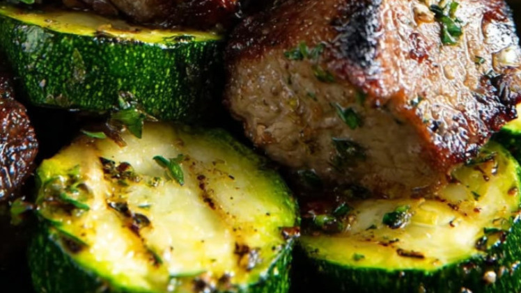 Cast iron skillet of golden-seared garlic butter steak bites plated alongside caramelized zucchini, garnished with fresh parsley on a dark wood surface