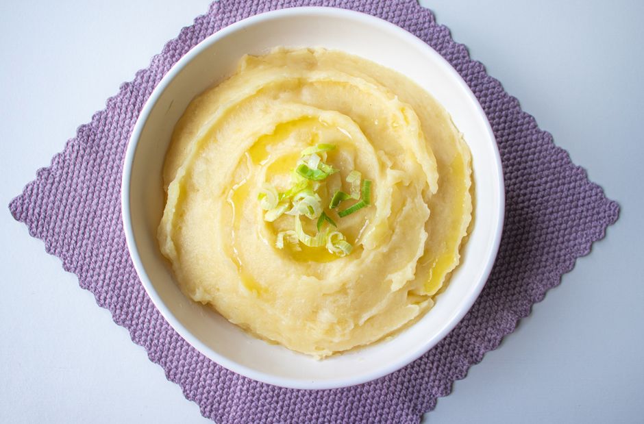 A white ceramic bowl of creamy homemade mashed potatoes topped with a pat of melting butter and a sprinkle of fresh chives, set on a wooden table with a linen napkin