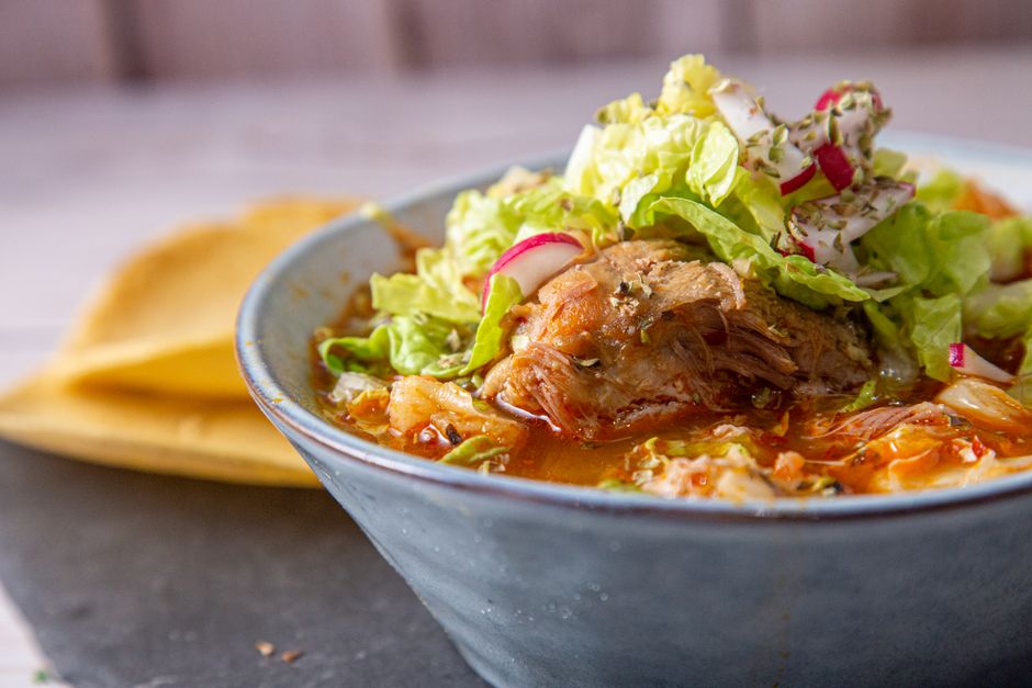 A deep bowl of red pozole rojo topped with shredded cabbage, sliced radishes, diced onion, and a lime wedge, with tostadas on the side