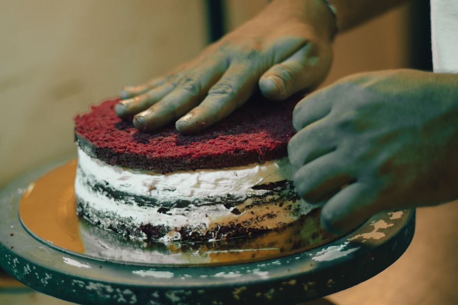Tall two-layer red velvet cake with white cream cheese frosting on a white cake stand, one slice removed to show the deep red interior crumb