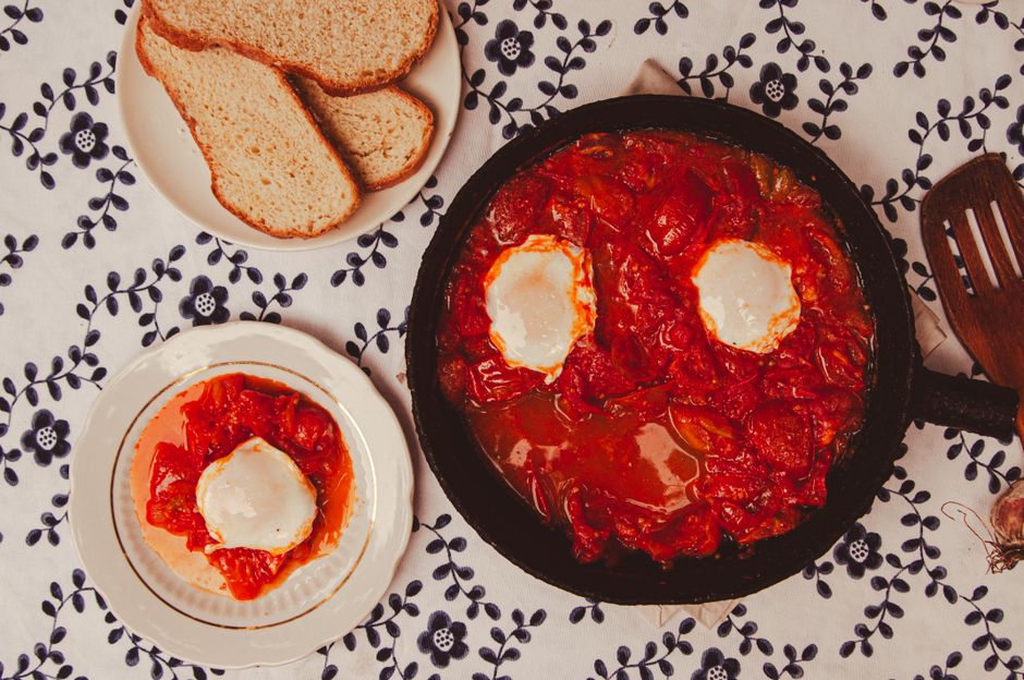 Shakshuka in a cast iron skillet with six poached eggs nestled in rich red spiced tomato and pepper sauce, topped with crumbled feta and chopped parsley