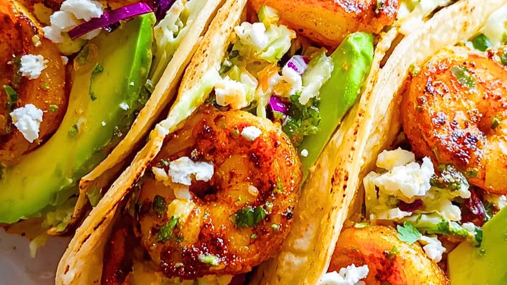 Close-up of two shrimp tacos on corn tortillas topped with cabbage slaw, sliced avocado, chipotle crema drizzle, and fresh cilantro on a wooden board with lime wedges