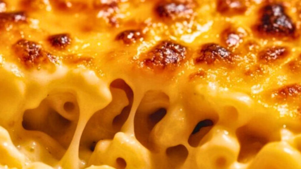 A close-up of a golden-topped Southern baked mac and cheese in a white baking dish, with a wooden spoon lifting a creamy, cheese-pulling scoop from the center