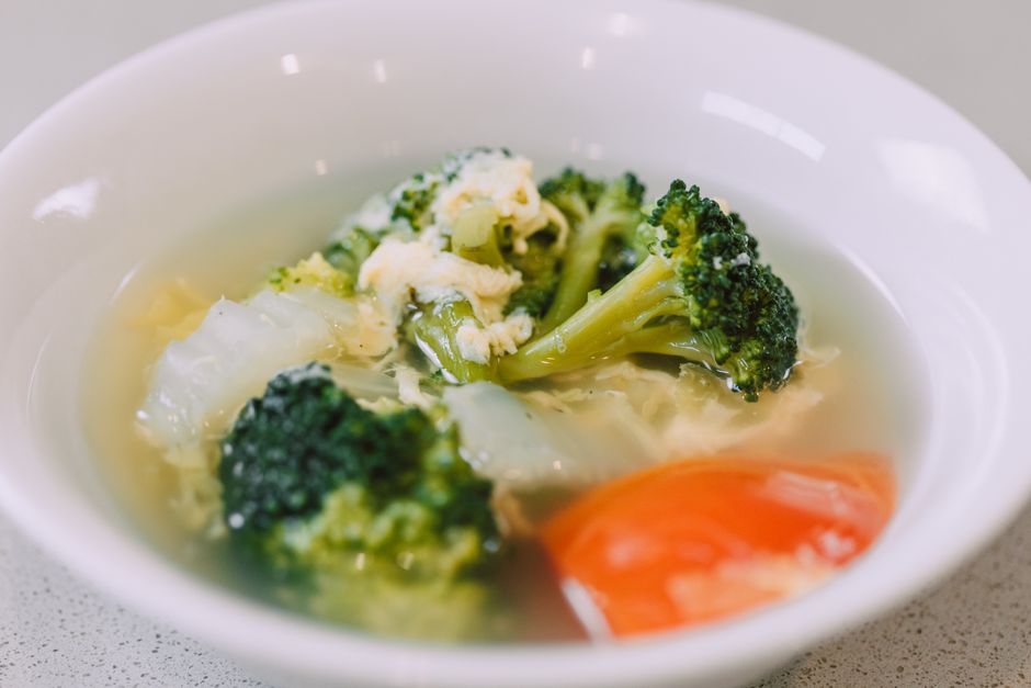 A white ceramic bowl of hearty homemade vegetable soup filled with colorful carrots, potatoes, green beans, zucchini, and spinach in a rich tomato broth, photographed from above on a wooden table with crusty bread alongside