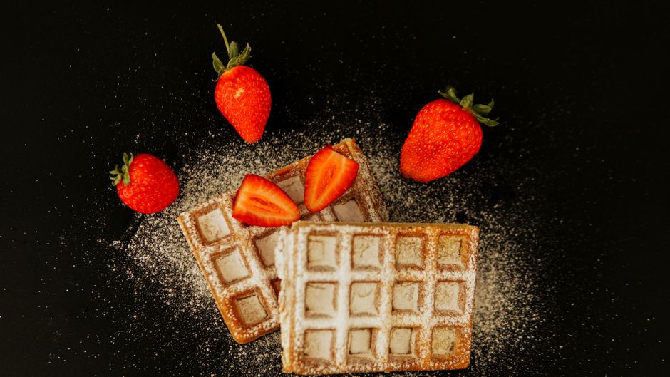 Stack of golden crispy homemade waffles on a white plate with fresh berries and maple syrup drizzled over the top, photographed from above on a light wood surface